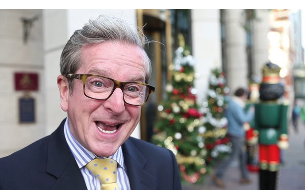 A person wearing glasses, a suit, and a yellow tie is smiling in front of a decorated Christmas tree and a large nutcracker figure on a city street. Description generated by AI