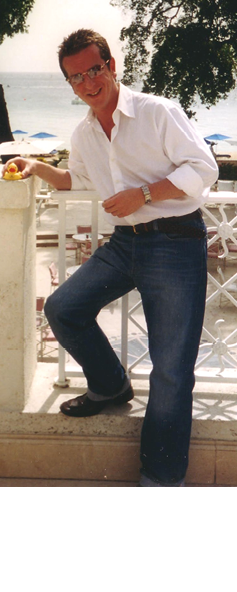 A person wearing glasses, a white shirt, and jeans is standing outdoors near a railing with trees and a beach visible in the background. Description generated by AI