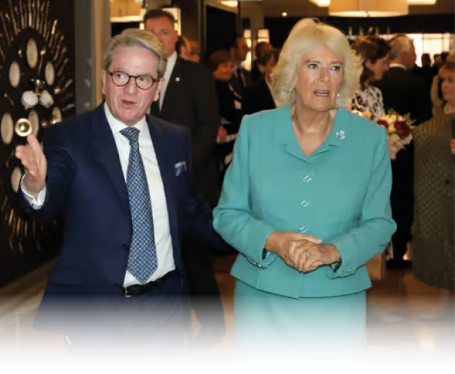 Two adults dressed in formal attire are engaged in conversation while walking indoors at a social event with other people in the background. Description generated by AI
