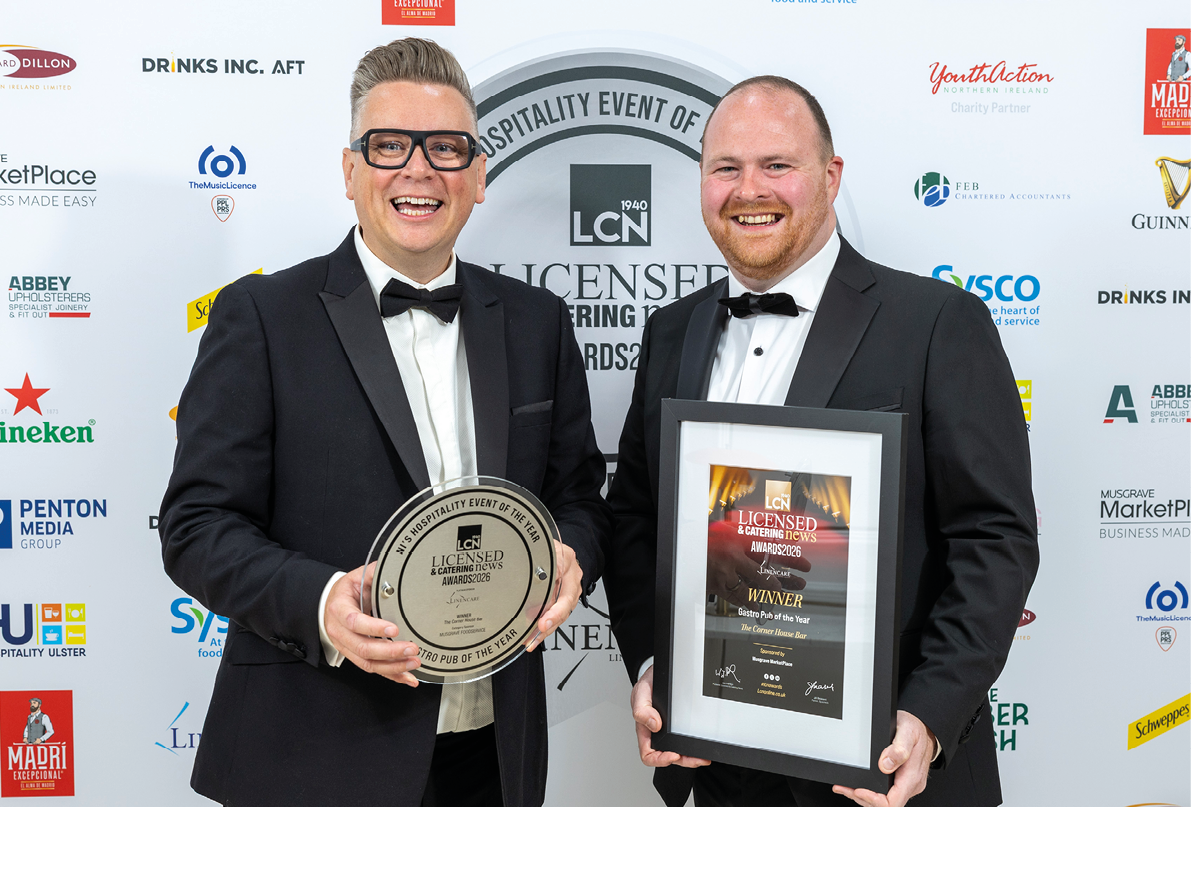 Two adults wearing tuxedos smile while holding an award plaque and a framed certificate at a formal event with a branded backdrop. Description generated by AI