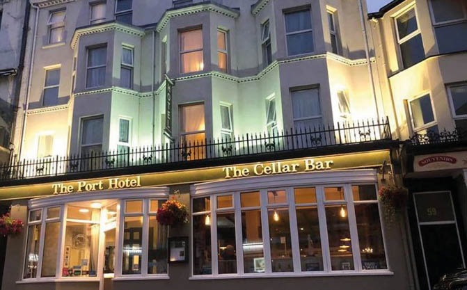 A warmly lit building exterior at night features The Port Hotel and The Cellar Bar with hanging flower baskets and large windows. Description generated by AI
