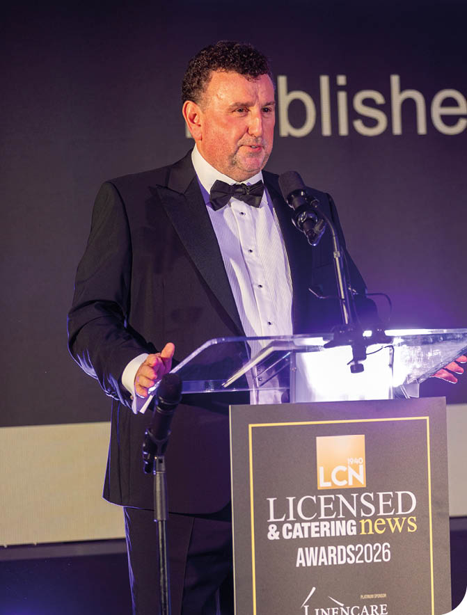 A person wearing a black suit jacket and white dress shirt is standing behind a clear podium with a microphone. Description generated by AI