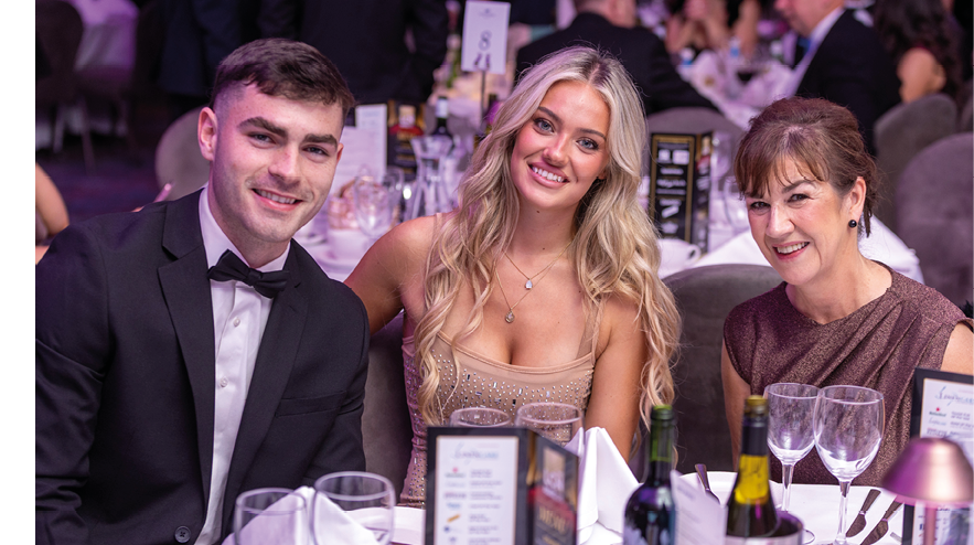 Three adults dressed in formal attire are sitting closely together at a decorated table with wine glasses and menus, smiling in a banquet or event setting. Description generated by AI