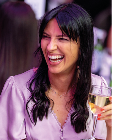 A person with long dark hair wearing a light purple blouse is laughing and holding a glass of white wine in a lively social setting. Description generated by AI