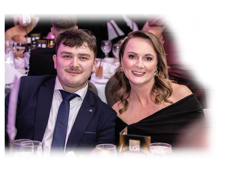 Two adults dressed formally are sitting closely together at a table with glasses and a menu card in a dimly lit event setting. Description generated by AI
