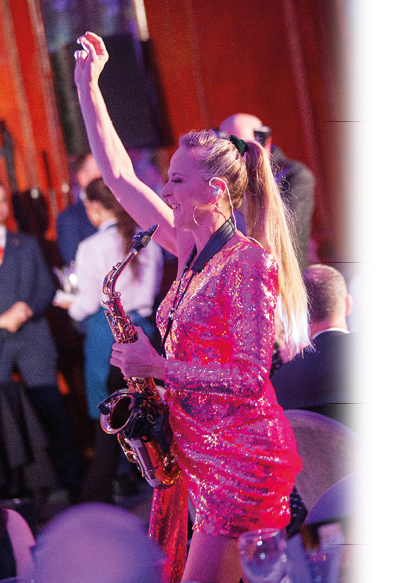 A person with long hair tied in a ponytail is wearing a sparkly red dress and playing a saxophone while raising one arm in a lively indoor setting with other people in the background. Description generated by AI