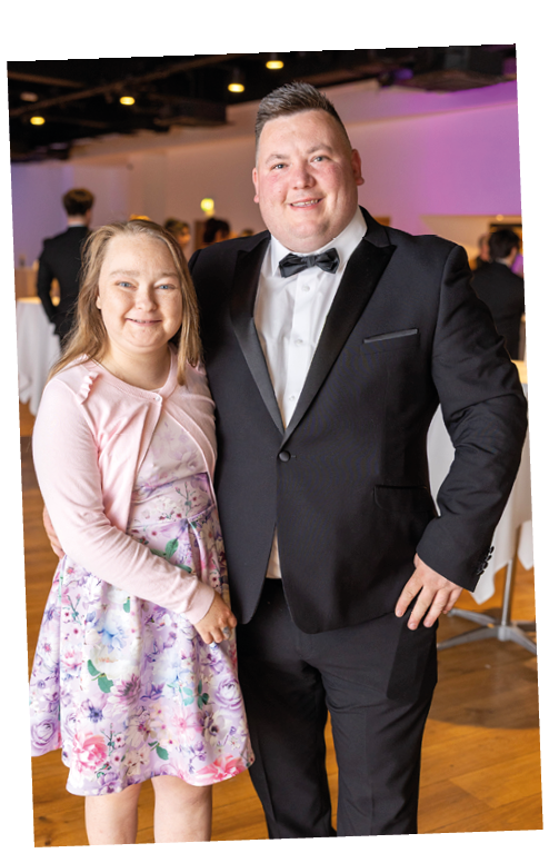 Two adults dressed formally with one wearing a black tuxedo and bow tie and the other wearing a floral dress and light pink cardigan, standing closely together and smiling indoors. Description generated by AI
