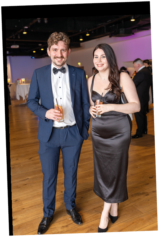 Two adults dressed in formal attire are standing indoors on a wooden floor, each holding a drink and smiling. Description generated by AI