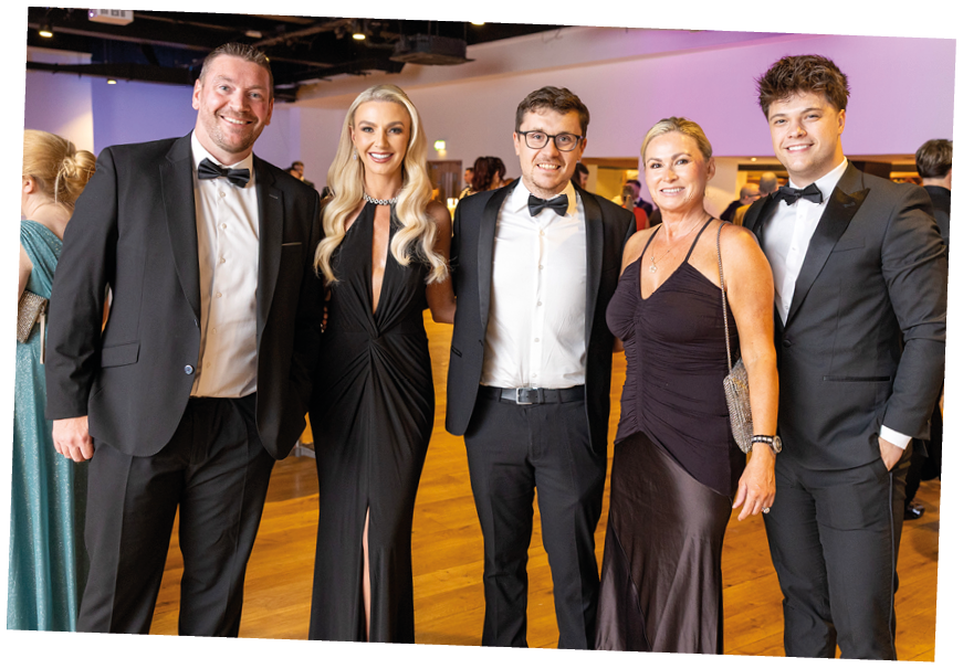 Five adults dressed in formal evening attire stand closely together indoors with a wooden floor and soft lighting in the background. Description generated by AI