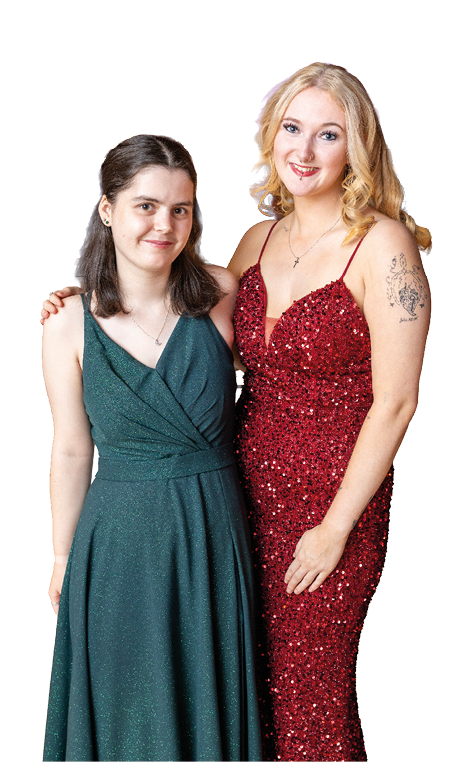 Two adults stand closely together smiling, one wearing a dark green sleeveless dress and the other wearing a red sequined dress with a tattoo on the upper arm. Description generated by AI