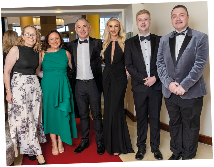 Six adults dressed in formal attire stand closely together on a red carpet in an indoor setting, smiling and posing for the photo. Description generated by AI