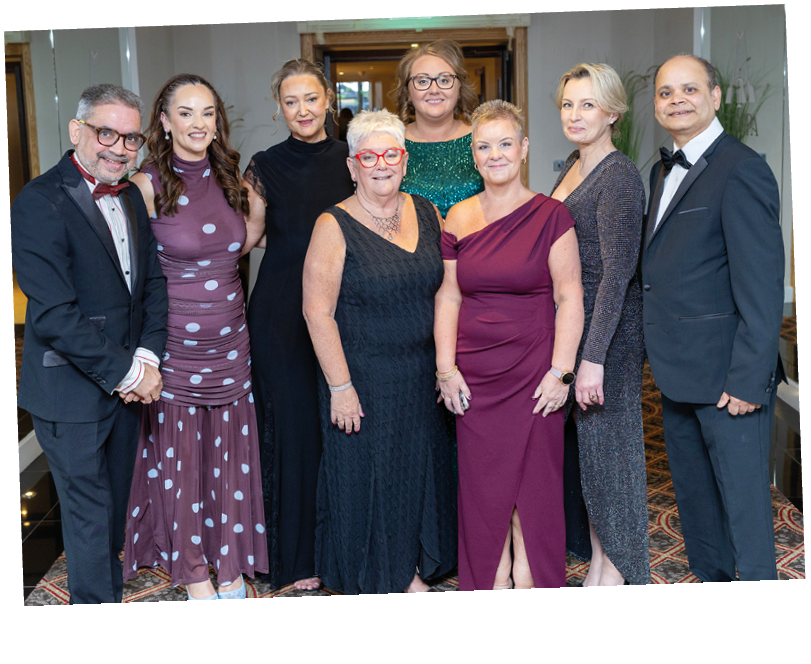 A group of eight adults dressed in formal attire stand closely together indoors, smiling and posing for a photo. Description generated by AI