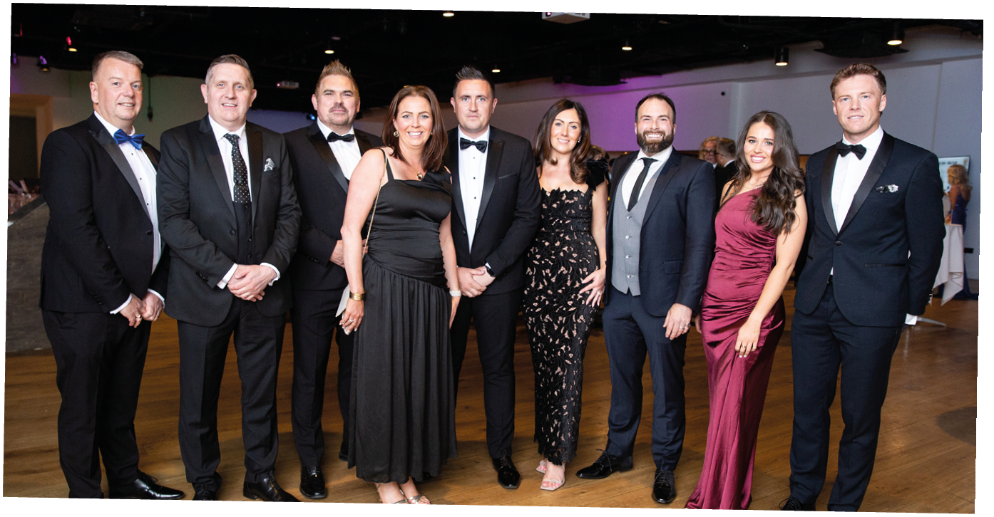 A group of adults dressed in formal evening attire stand together on a wooden floor in a well-lit indoor event space. Description generated by AI
