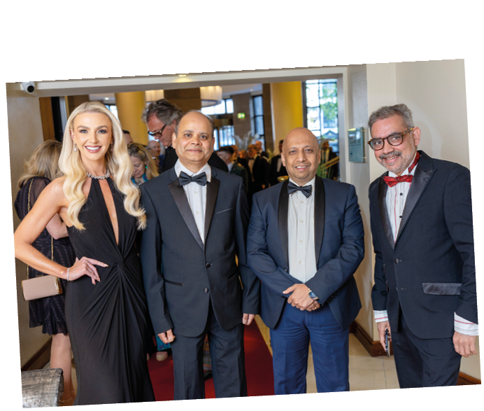 Four adults dressed in formal attire stand together indoors on a red carpet with a crowd in the background. Description generated by AI