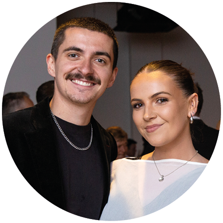 Two adults stand close together indoors, one wearing a black shirt and chain necklace, and the other wearing a white top with a pendant necklace. Description generated by AI