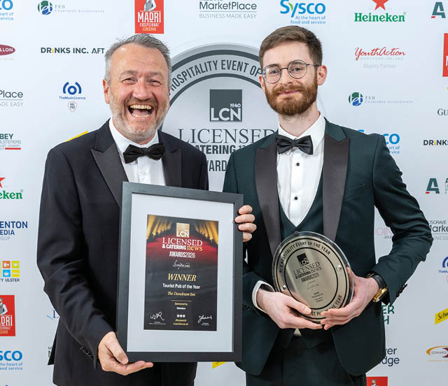 Two adults dressed in formal suits and bow ties smile while holding framed awards at a hospitality event. Description generated by AI