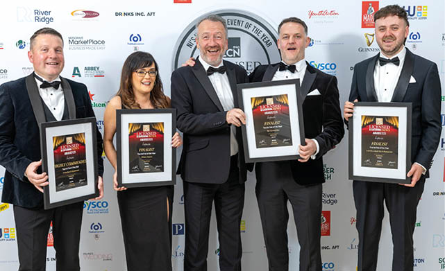 Five adults dressed in formal attire stand in front of a backdrop holding framed awards and smiling. Description generated by AI