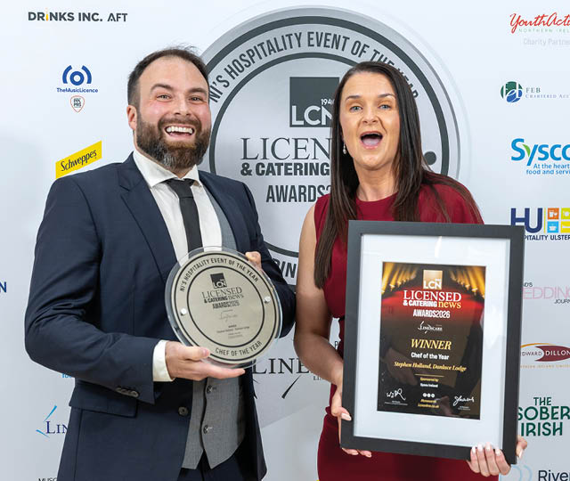 Two adults dressed in formal attire are smiling and holding framed awards at a hospitality event with a branded backdrop. Description generated by AI