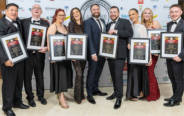 A group of adults dressed in formal attire stand smiling and holding framed awards in front of a backdrop with various logos. Description generated by AI