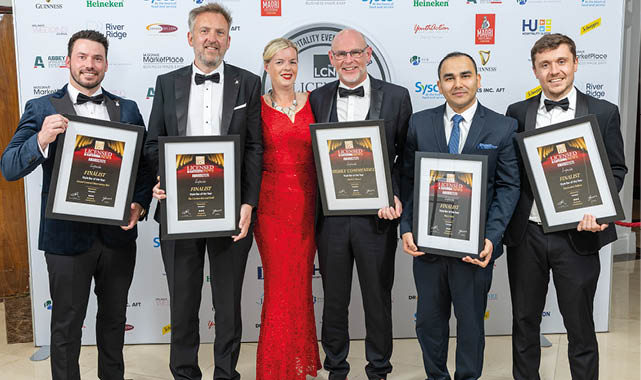 Five adults dressed in formal attire stand side by side holding framed awards against a backdrop with logos. Description generated by AI