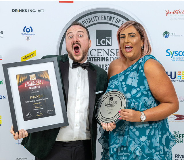 Two adults dressed formally are smiling and holding framed awards at a hospitality event with sponsor logos in the background. Description generated by AI