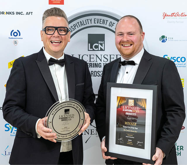 Two adults in tuxedos smiling and holding awards at a hospitality event. Description generated by AI