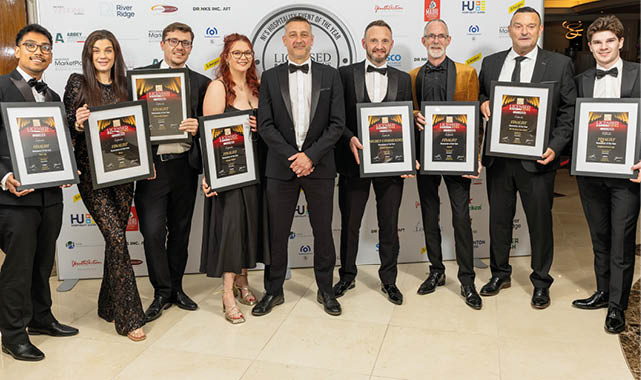 A group of eight adults dressed in formal attire stand in a row holding framed awards in front of a backdrop with various logos. Description generated by AI