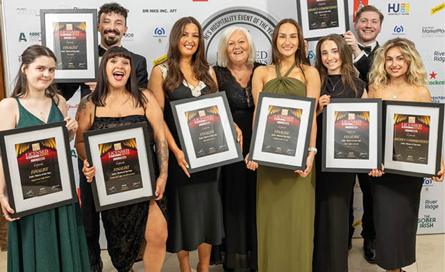 A group of adults dressed in formal attire stand together smiling and holding framed awards at an event with a branded backdrop. Description generated by AI