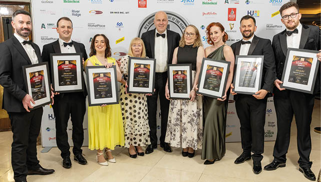 A group of eight adults dressed in formal attire stand side by side holding framed awards in front of a backdrop covered with various sponsor logos. Description generated by AI
