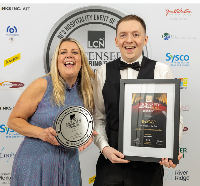Two adults dressed in formal attire smile while holding an award plaque and a framed certificate at a hospitality event. Description generated by AI