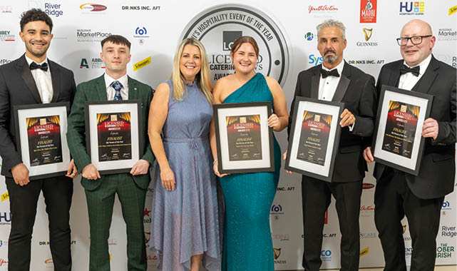 Five adults dressed in formal attire stand smiling in front of a branded backdrop, with four holding framed awards. Description generated by AI
