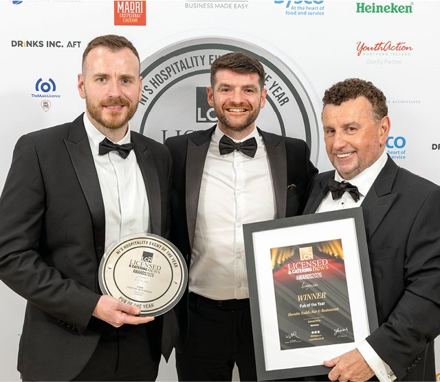 Three adults dressed in tuxedos stand together smiling, with one holding a framed award and another holding a round plaque at an event backdrop. Description generated by AI