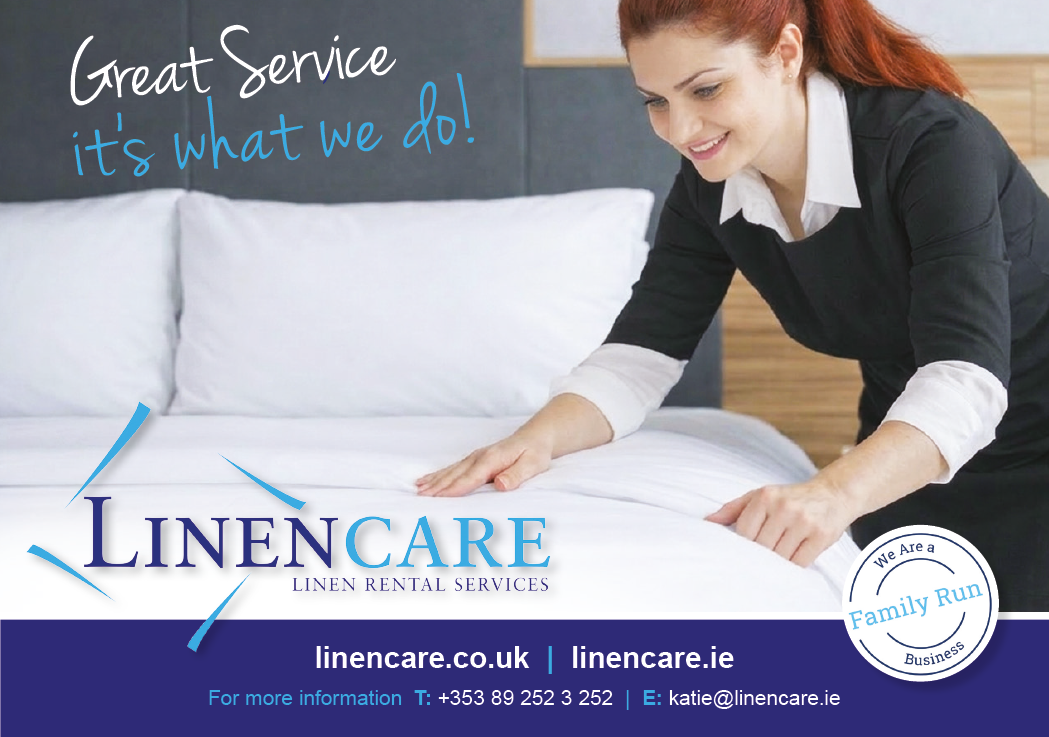 A person with long hair tied back wearing a black sweater over a white collared shirt is making a bed in a modern bedroom with a promotional text for linen care services visible on the image. Description generated by AI