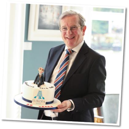 A person wearing a suit and striped tie is smiling and holding a cake decorated with a small figurine and the word Europa in a bright room with a window and framed artwork in the background. Description generated by AI