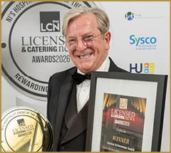 A senior person wearing a tuxedo is smiling while holding a framed award certificate and a round gold trophy at a hospitality event. Description generated by AI