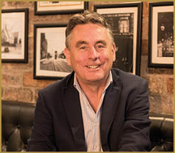 A person wearing a dark blazer, light checkered shirt, and jeans is sitting cross-legged on a black leather couch in a room with brick walls and framed black-and-white photographs. Description generated by AI