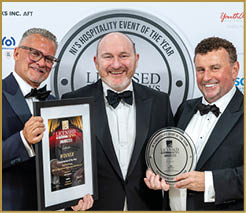 Three adults dressed in formal tuxedos smile while holding framed awards at a hospitality event with a branded backdrop. Description generated by AI
