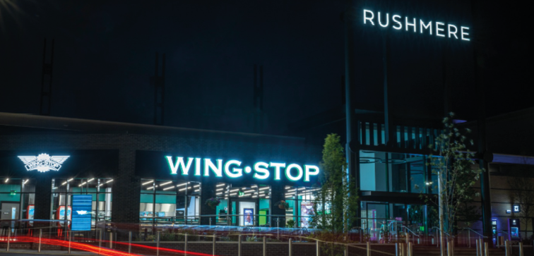 A Wingstop restaurant with illuminated signage is shown at night with light trails from passing vehicles and a nearby sign reading Rushmere. Description generated by AI