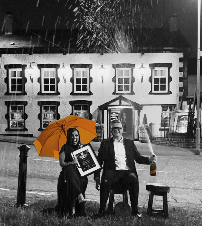 Two people sit on chairs on grass at night in front of a building with one holding an orange umbrella and the other holding a bottle and a framed picture while sparks fly above them. Description generated by AI