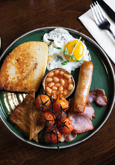 A plate with a cooked breakfast including toast, fried egg, baked beans, sausage, grilled tomatoes, bacon, and a piece of fried bread on a wooden table next to a fork and knife on a napkin. Description generated by AI