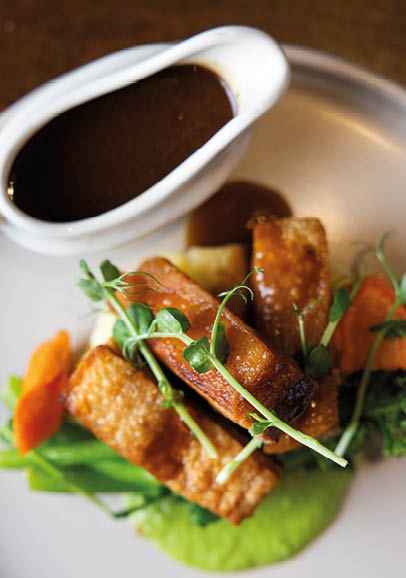 A gourmet dish featuring crispy pork belly with green vegetables, mashed peas, and a side of rich brown sauce in a white gravy boat. Description generated by AI