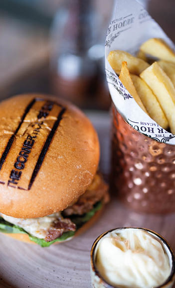 A burger with lettuce and melted cheese on a branded bun is served on a plate with a copper cup holding fries wrapped in paper and a small bowl of creamy dipping sauce. Description generated by AI