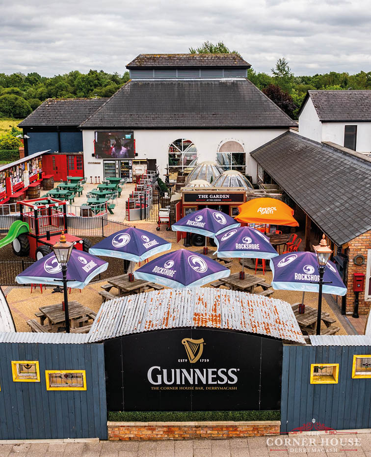 An outdoor pub garden with multiple tables shaded by branded umbrellas, a playground slide, and a large building with a Guinness sign on the fence in front. Description generated by AI