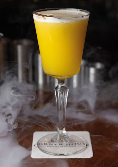 A yellow cocktail with foam on top is served in a tall glass on a coaster with smoke swirling around it on a wooden bar counter. Description generated by AI
