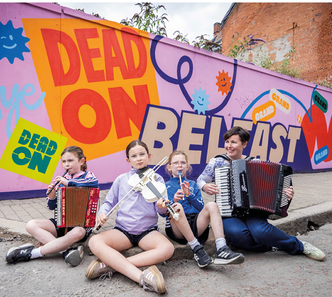 Four people sitting on the ground in front of a colorful mural playing musical instruments including accordions and a violin. Description generated by AI