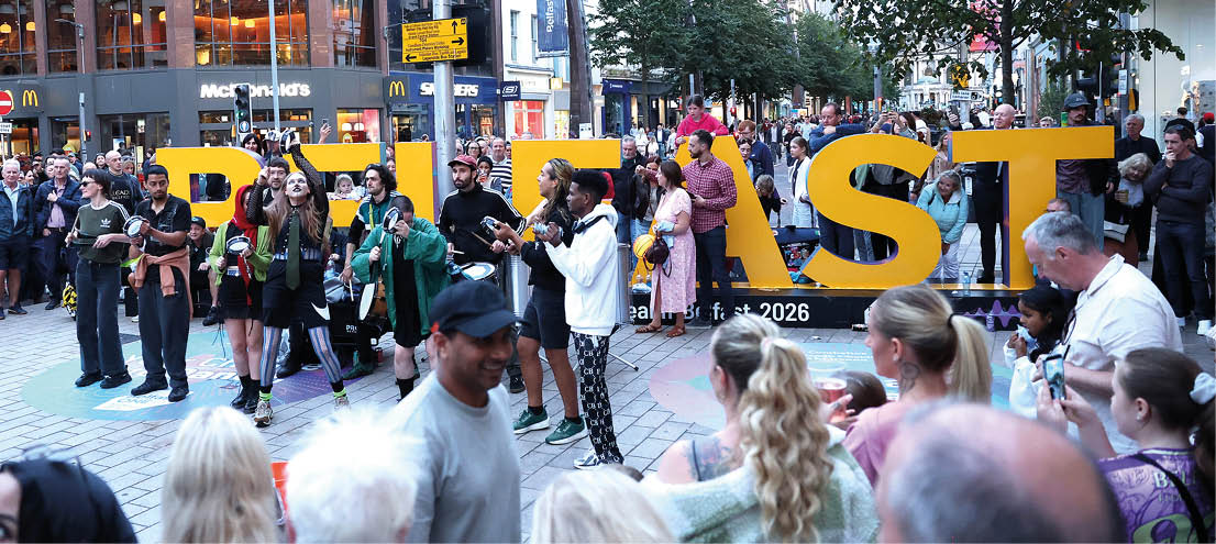 A crowd of people gathers outdoors around large yellow letters spelling “RUTST" with some individuals taking photos and others watching the scene in an urban setting. Description generated by AI