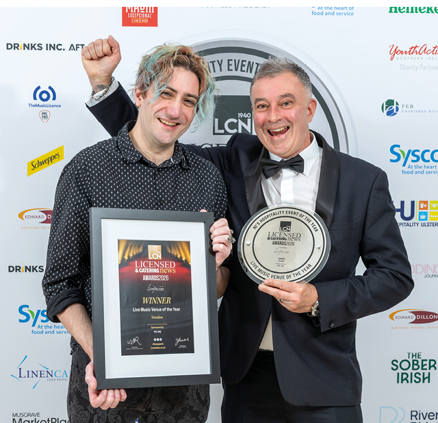 Two adults smiling and celebrating while holding an award plaque and a round award at a formal event with a branded backdrop. Description generated by AI