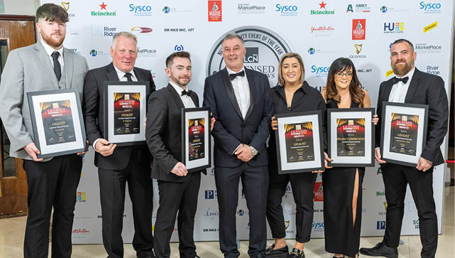 Seven adults dressed in formal attire stand in a row holding framed awards against a backdrop with logos. Description generated by AI