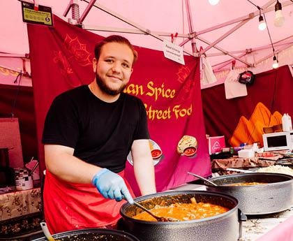 A person wearing a black shirt, red apron, and blue gloves is serving food from large pots at a colorful outdoor food stall decorated with string lights and a pink banner. Description generated by AI