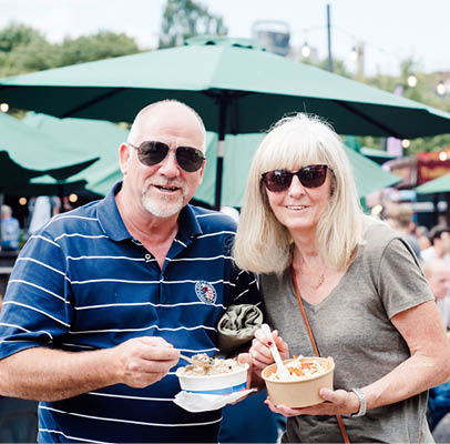 Two adults wearing sunglasses are smiling and holding bowls of food while standing outdoors at a busy market or festival with green umbrellas and string lights in the background. Description generated by AI